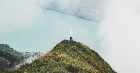 Adventure - A Person Standing on a Grassy Cliff near the Clouds