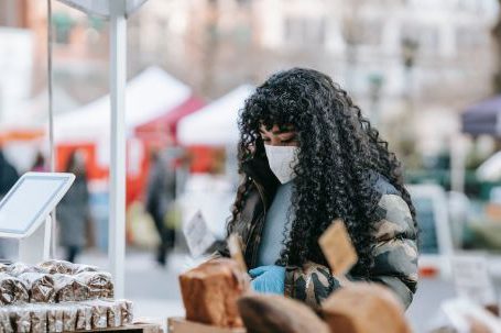 Local Fairs - Young African American female in warm casual outfit and medical mask looking at showcase with various breads at street fair