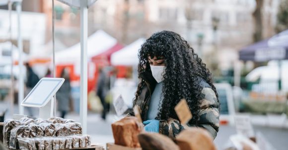 Local Fairs - Young African American female in warm casual outfit and medical mask looking at showcase with various breads at street fair