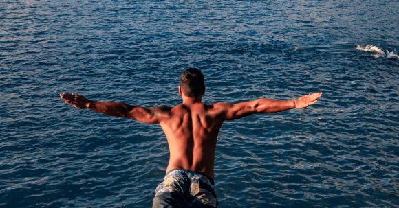Wellness Travel - Back view of unrecognizable shirtless muscular male traveler jumping into blue sea water with outstretched arms on sunny day