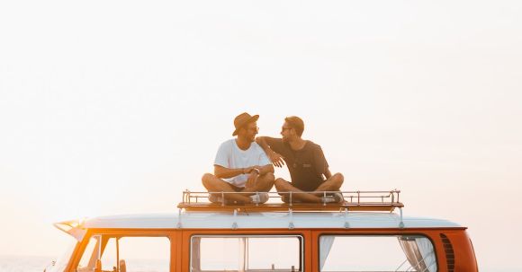 Road Trip - Silhouettes of full body faceless male friends in casual clothes resting on camper van roof and chatting in countryside in sunset time