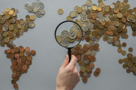 Travel Money - Anonymous person with magnifying glass over world map of coins