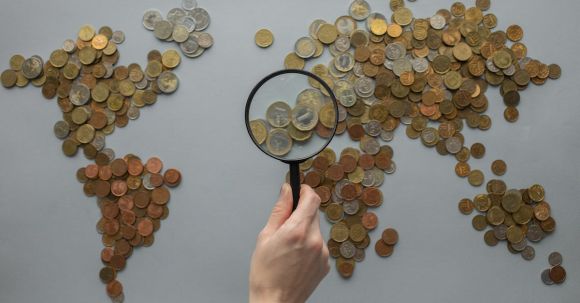 Travel Money - Anonymous person with magnifying glass over world map of coins