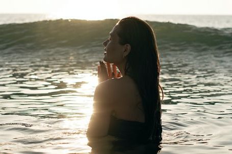 Travel Relaxation - Side view of tranquil young female tourist with long dark hair standing in waving ocean with closed eyes and enjoying summer sunset