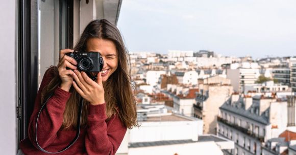 Sightseeing - Woman Wearing Maroon Sweater Standing on Veranda Using Camera While Smiling Overlooking Houses and Buildings