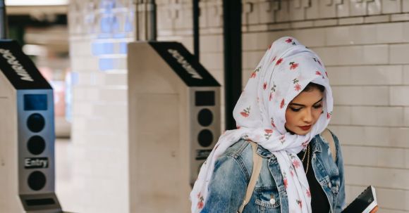 Independent Travel - Calm Muslim woman with copybook wearing floral headscarf wearing jeans jacket with backpack entering subway station through automatic turnstile