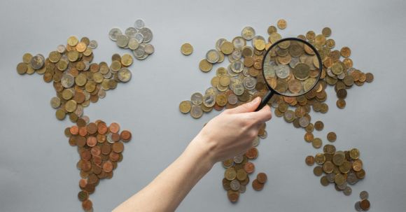 Travel Money - Top view of crop unrecognizable traveler with magnifying glass standing over world map made of various coins on gray background