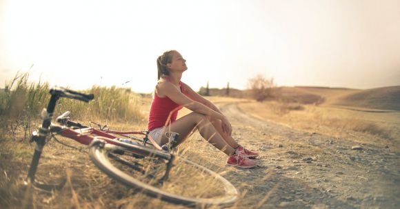Wellness Travel - Full body of female in shorts and top sitting on roadside in rural field with bicycle near and enjoying fresh air with eyes closed